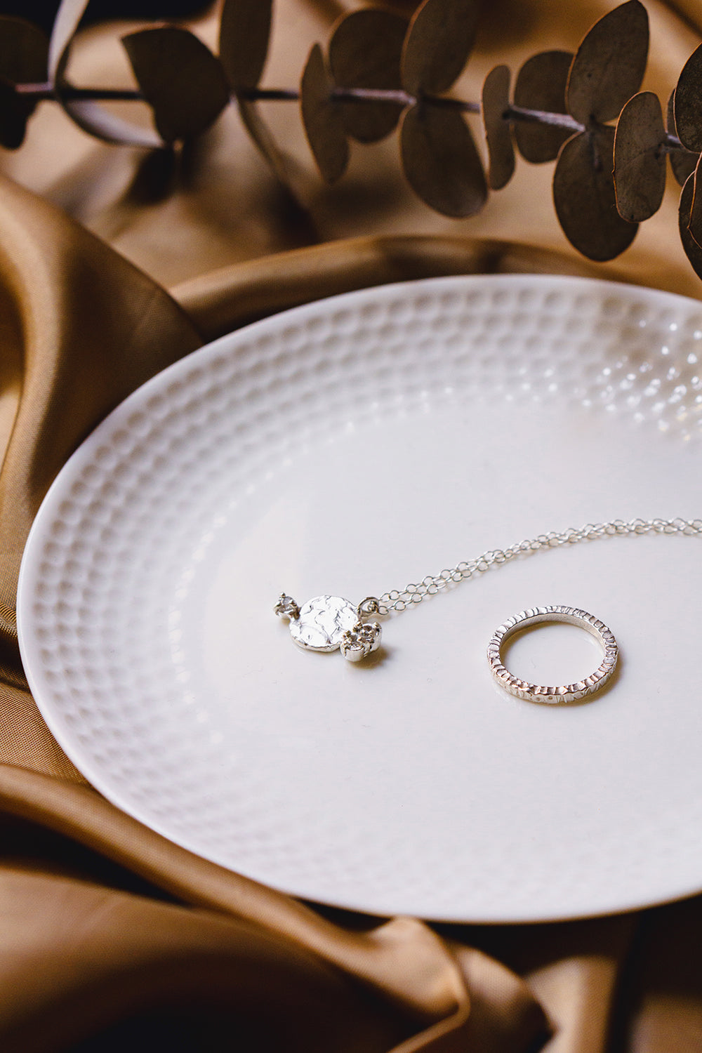 sterling silver textured round pendant necklace with white sapphires styled on flatlay with a textured silver ring on a white plate, surrounded by fabric and eucalyptus leaves